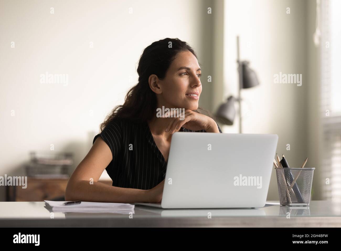 Smiling thoughtful female employee working at laptop from home Stock Photo
