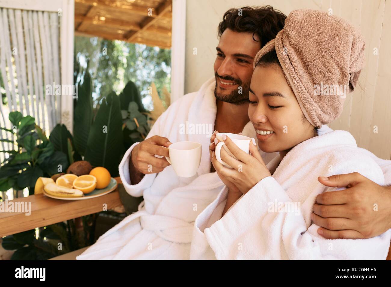 Beautiful multi-ethnic couple wearing white bathrobes drinking relaxing tea at spa. Romantic date at spa resort after relax and massage Stock Photo