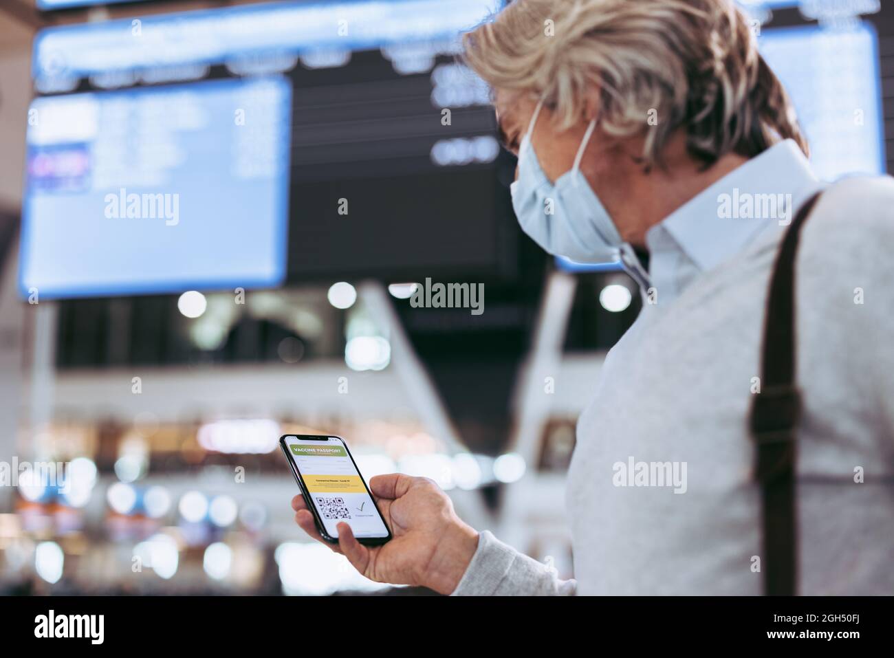 Male traveler holding his mobile phone with vaccine passport. Passenger wearing face mask at airport holding a smart phone with a digital certificate Stock Photo