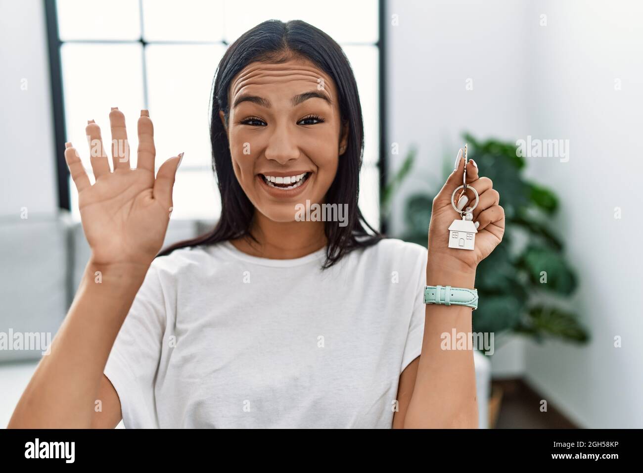 Young hispanic woman holding keys of new home waiving saying hello ...
