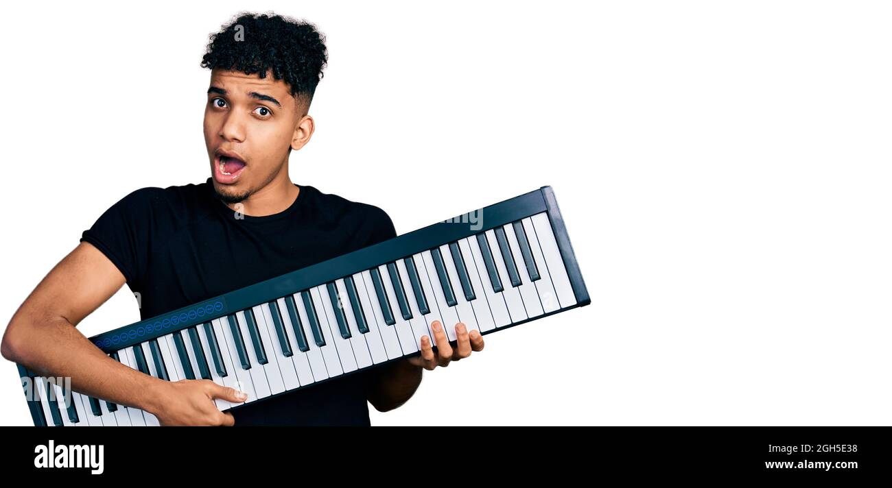 Young african american man holding piano keyboard in shock face, looking skeptical and sarcastic, surprised with open mouth Stock Photo