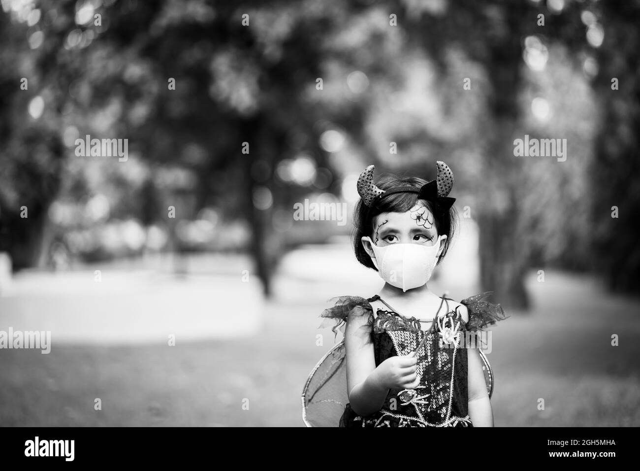 A grayscale shot of a Southeast Asian girl wearing her costume and also a facemask on Halloween, outdoors Stock Photo
