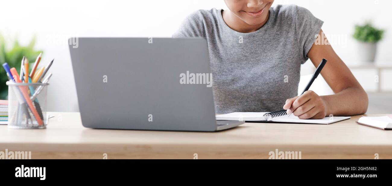 Cheerful adolescence afro american girl writes in notebook at laptop, studying at home Stock Photo