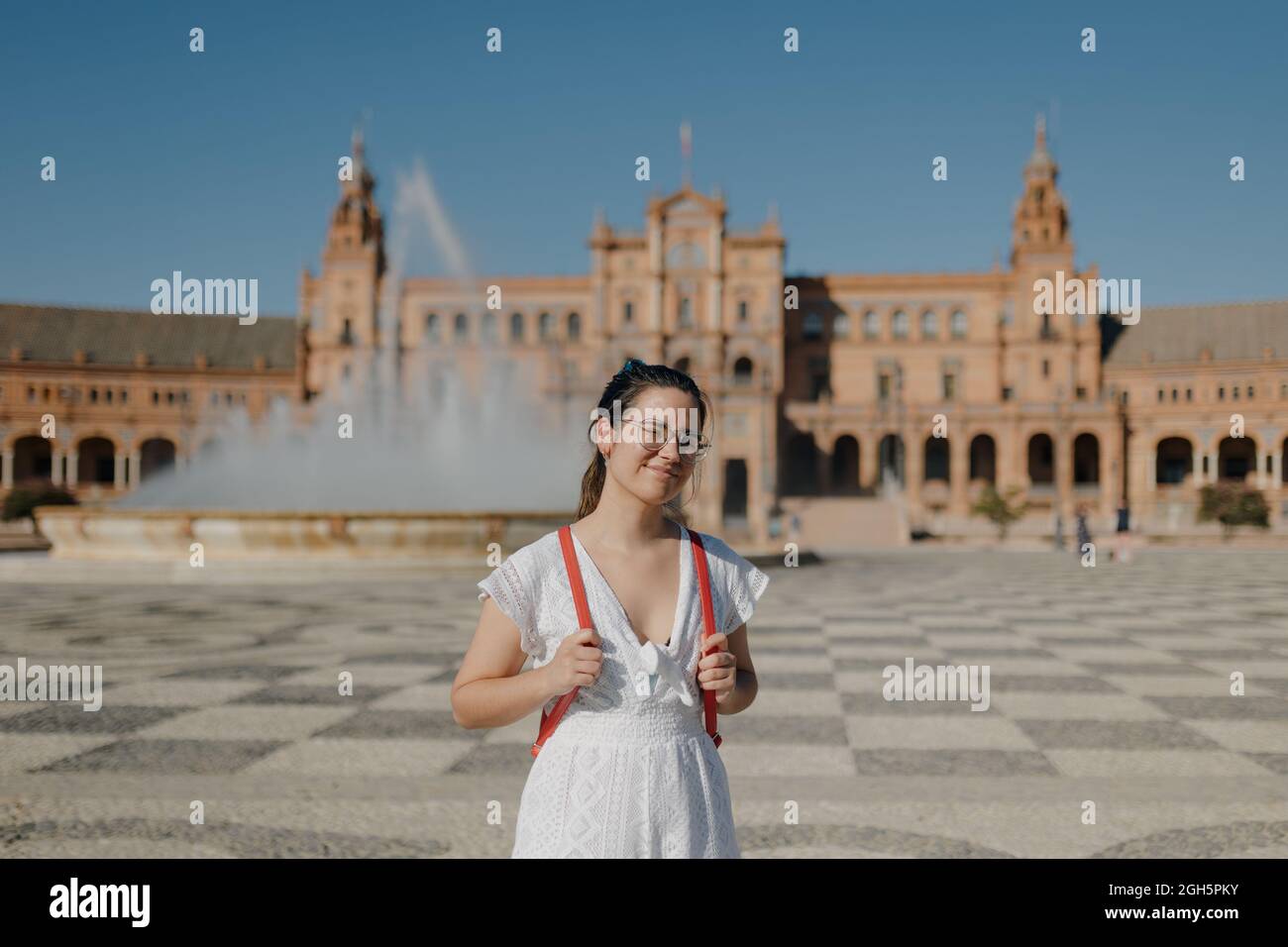 Young tourist woman with glasses wearing a white dress and red backpack smiles at camera while standing in the Plaza de España of Seville. Blinded by Stock Photo