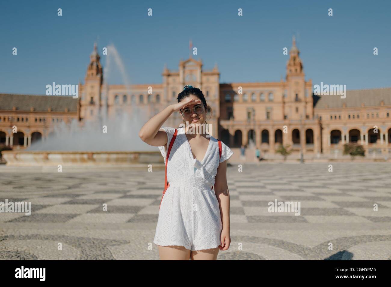 Young tourist woman with glasses wearing a white dress and red backpack is smiling and covering her eyes from the sun while standing in the Plaza de E Stock Photo