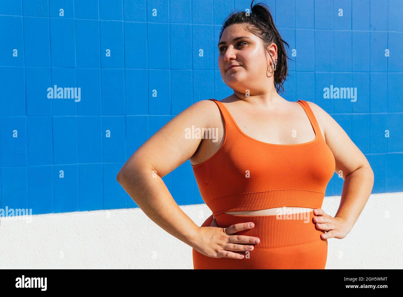 Overweight ethnic female athlete in active wear looking away against blue  tiled wall on sunny day Stock Photo - Alamy