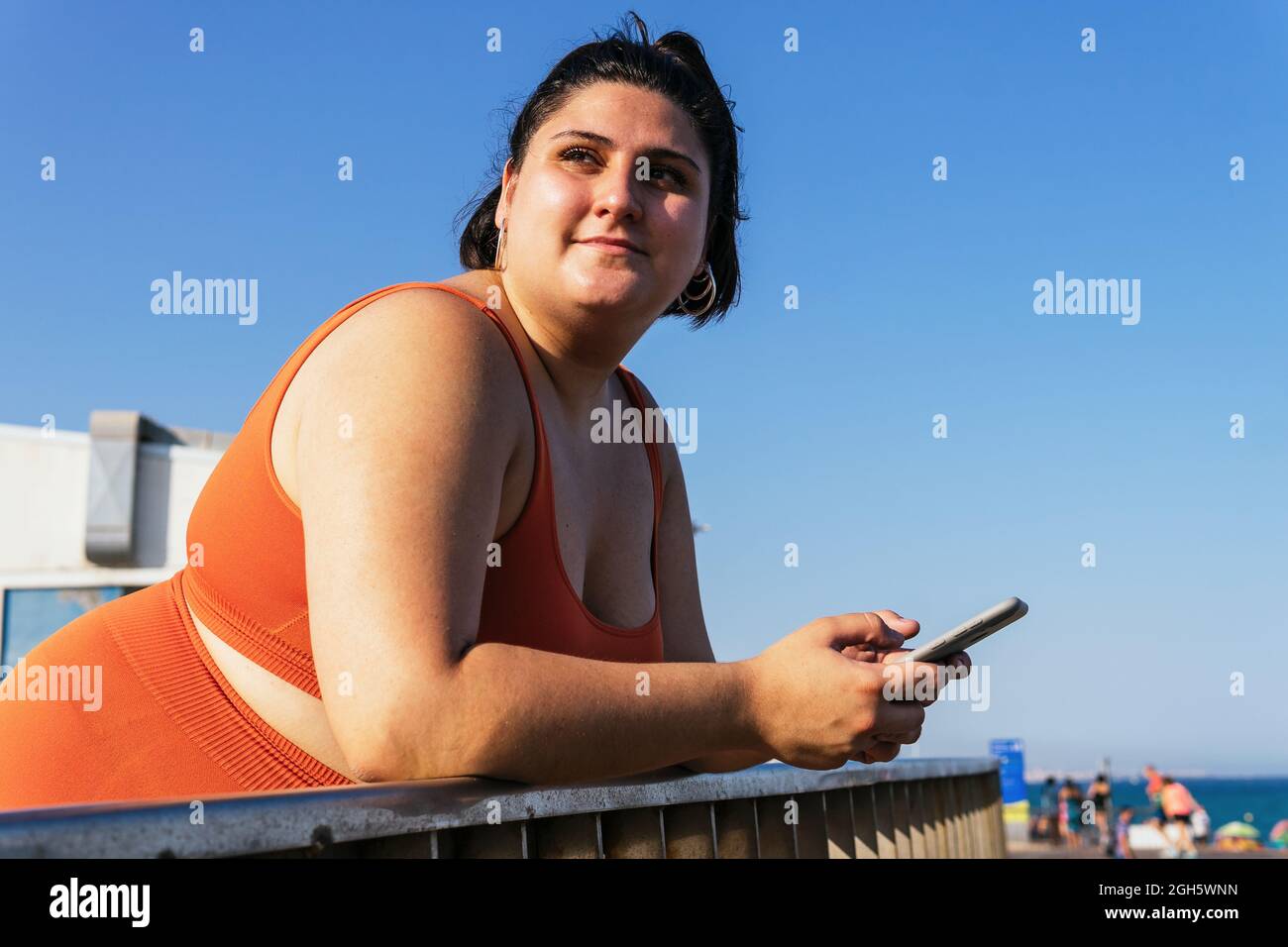 Dreamy ethnic female athlete with curvy body and cellphone leaning on fence while looking away under blue sky Stock Photo