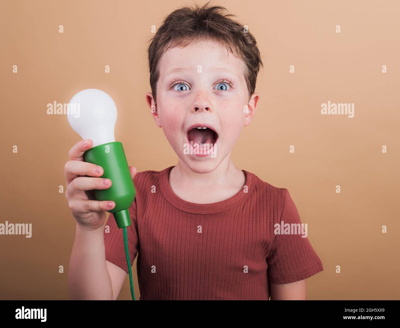 Pondering child in t shirt with plastic light bulb representing idea concept looking at camera on beige background Stock Photo