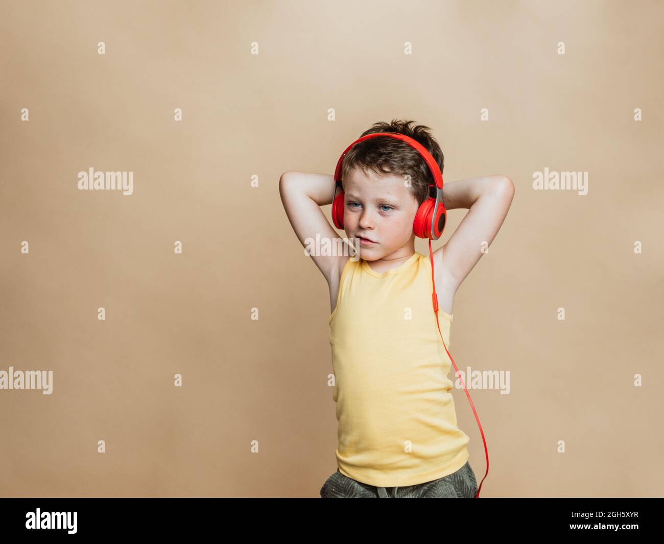 Carefree preteen boy in red headphones listening to music while standing on brown background and looking at camera Stock Photo