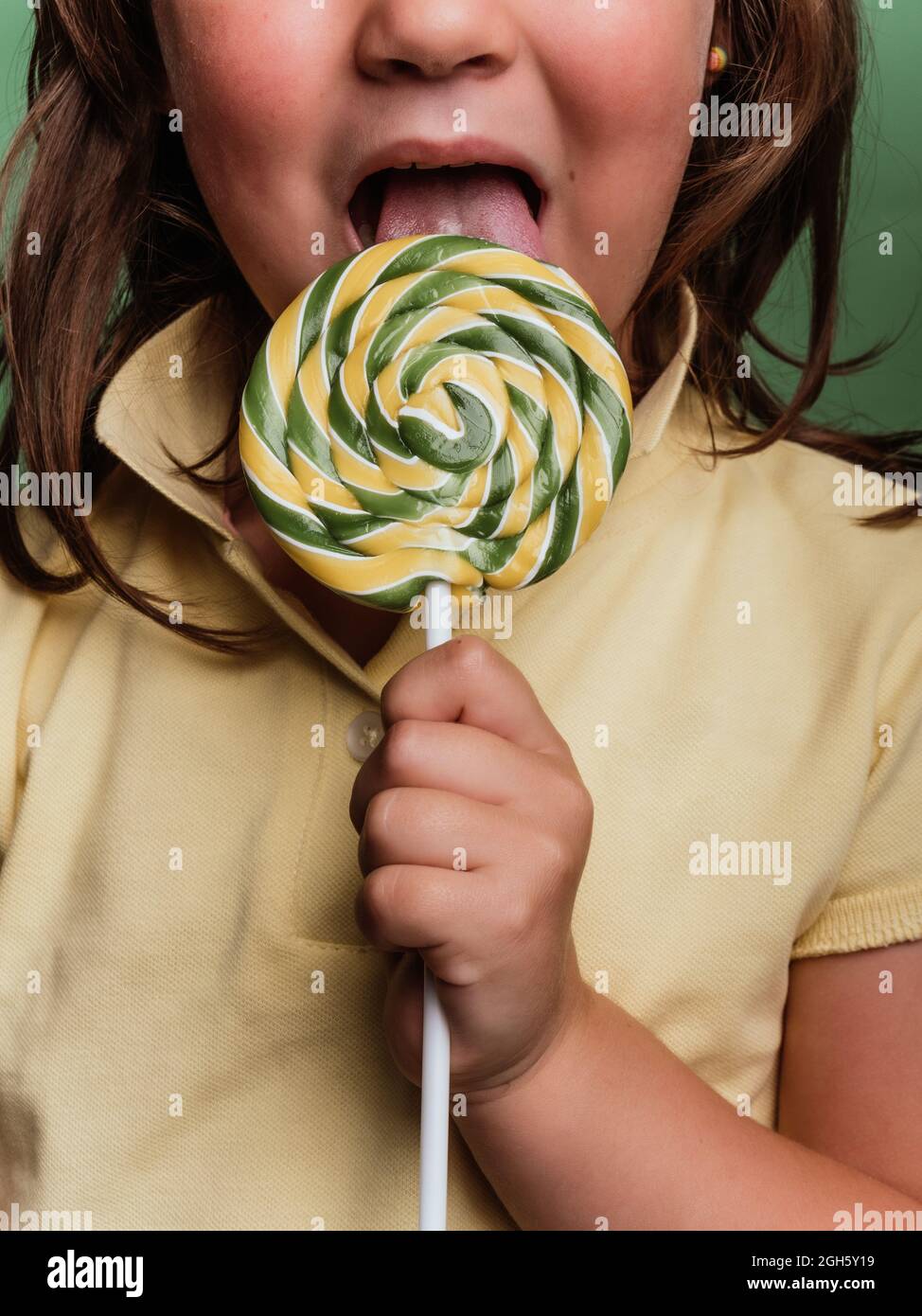 Anonymous kid reaching out hand with sweet swirl lollipop towards camera on green background in studio Stock Photo