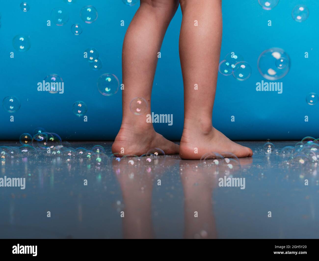 Crop barefoot child standing on floor in studio with flying soap bubbles on blue background Stock Photo