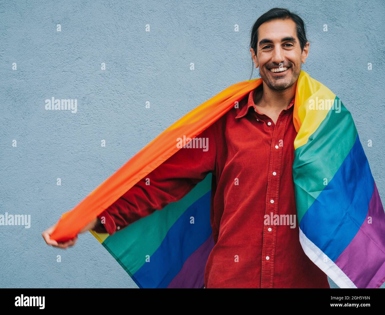 Gay ethnic male wrapped in rainbow LGBT flag looking at camera against ...
