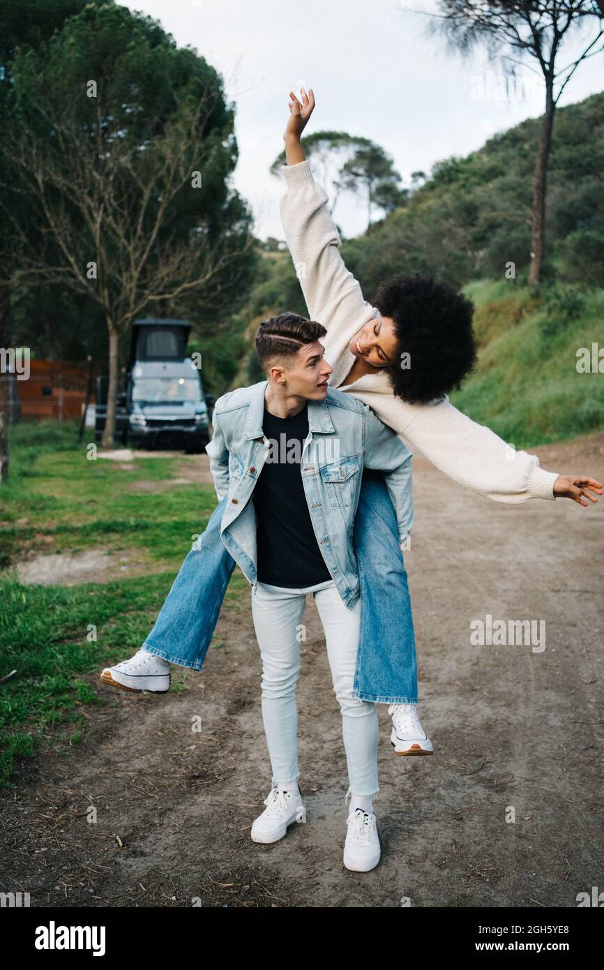 Young man giving cheerful ethnic girlfriend piggyback ride while looking away against camper and Thai Ridgeback on pathway Stock Photo