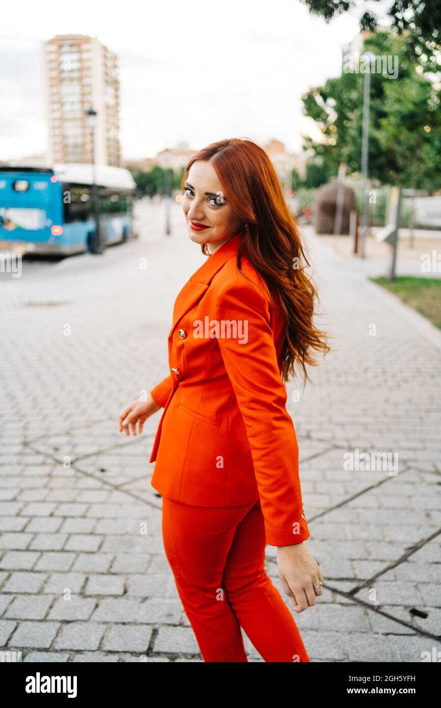 Back view of confident female with long ginger hair and in fashionable vivid red suit walking in city and looking at camera Stock Photo