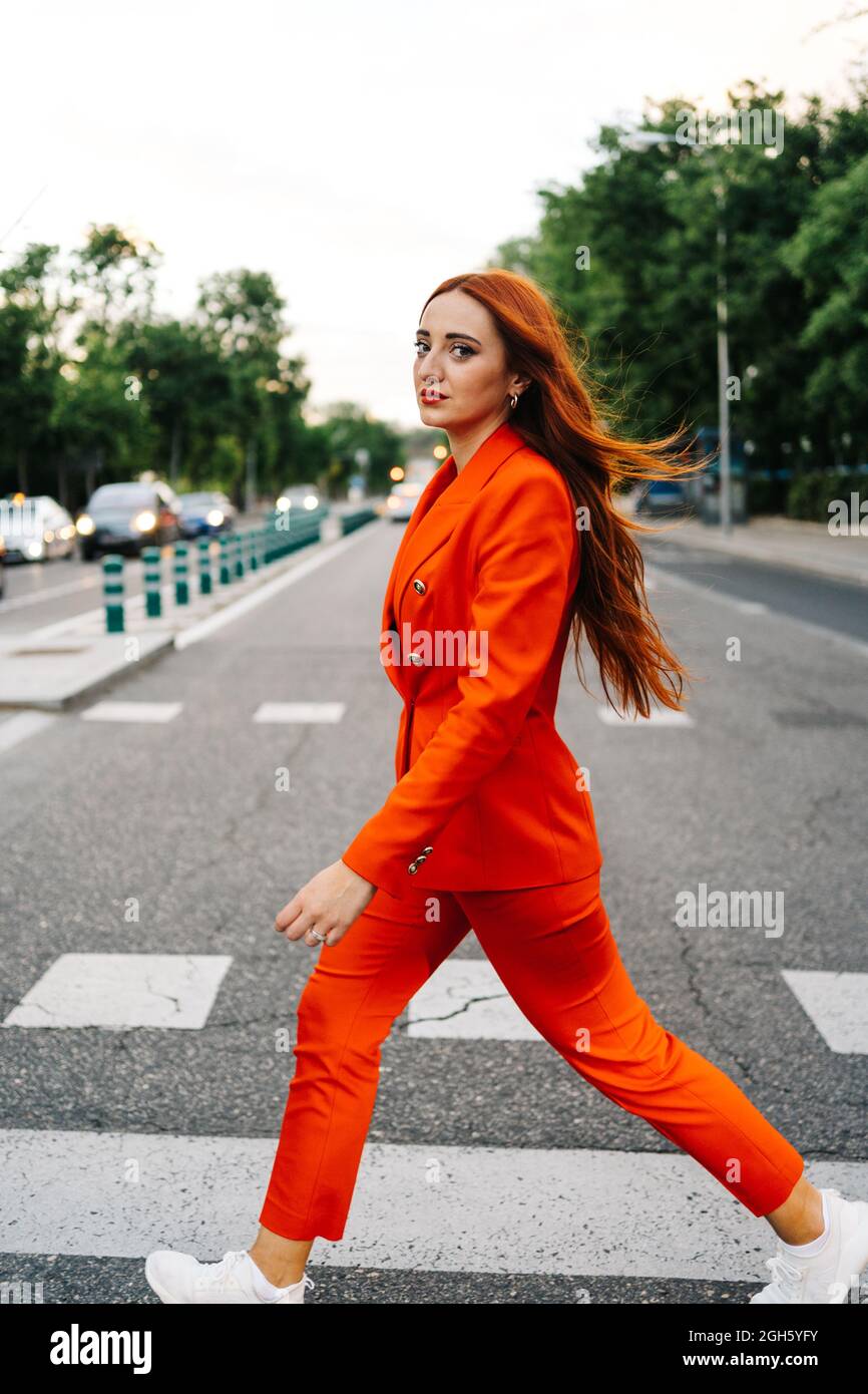 Side view of confident female with long ginger hair and in fashionable vivid orange suit crossing road in city and looking at camera Stock Photo