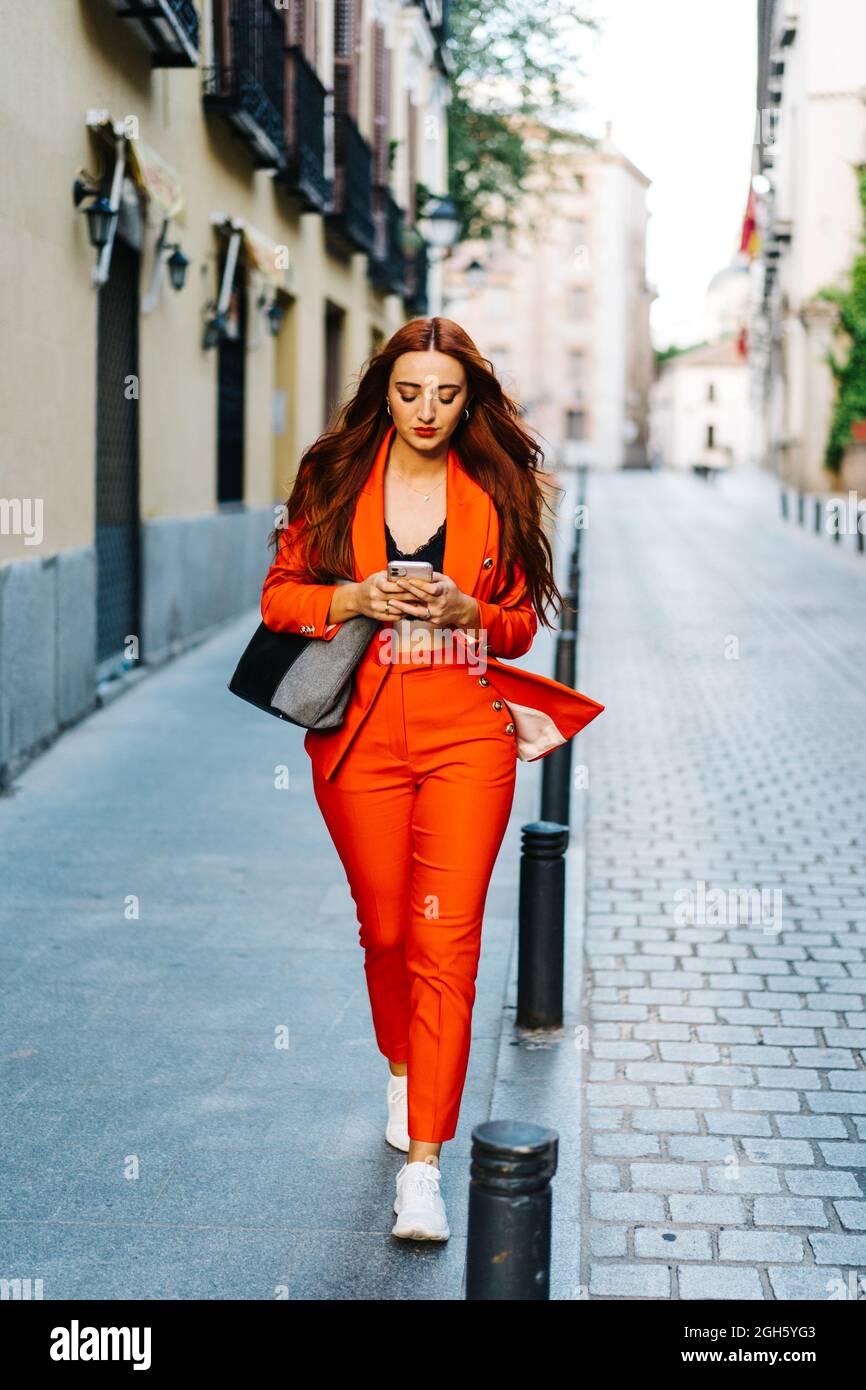 Stylish female with red hair and in vibrant red suit text messaging on smartphone in city street Stock Photo