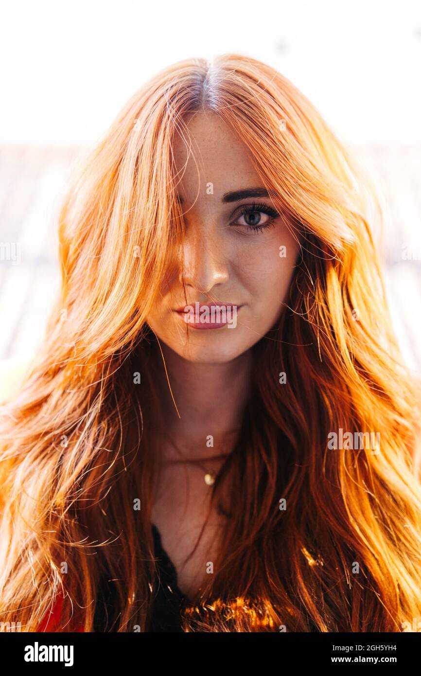 Pretty female with long red hair lit by sunlight looking at camera on blurred background Stock Photo