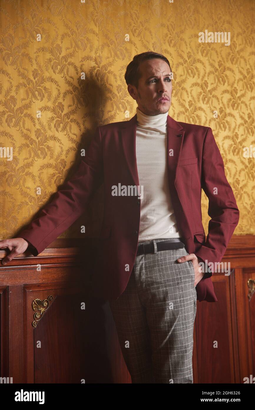 Confident adult male actor in elegant classy clothes keeping hand in pocket and looking away thoughtfully while standing near wall in vintage style ro Stock Photo