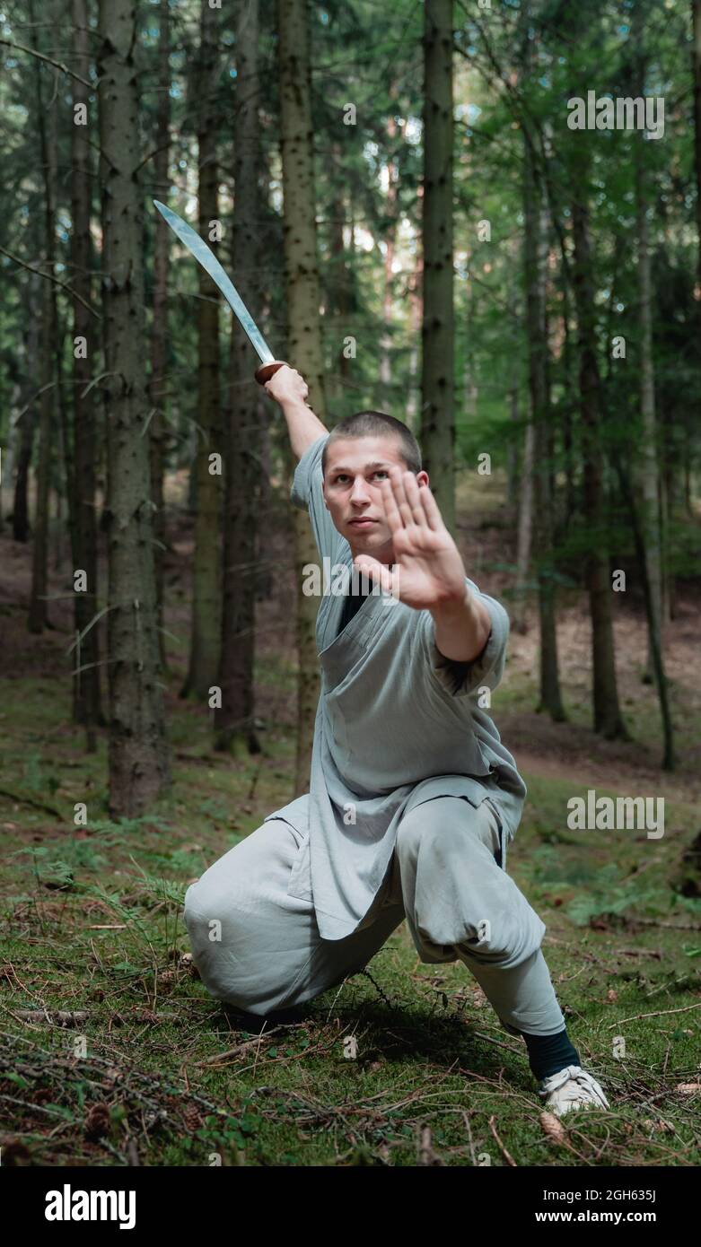 Full body man in traditional clothes practicing sword stance during kung fu training in forest Stock Photo