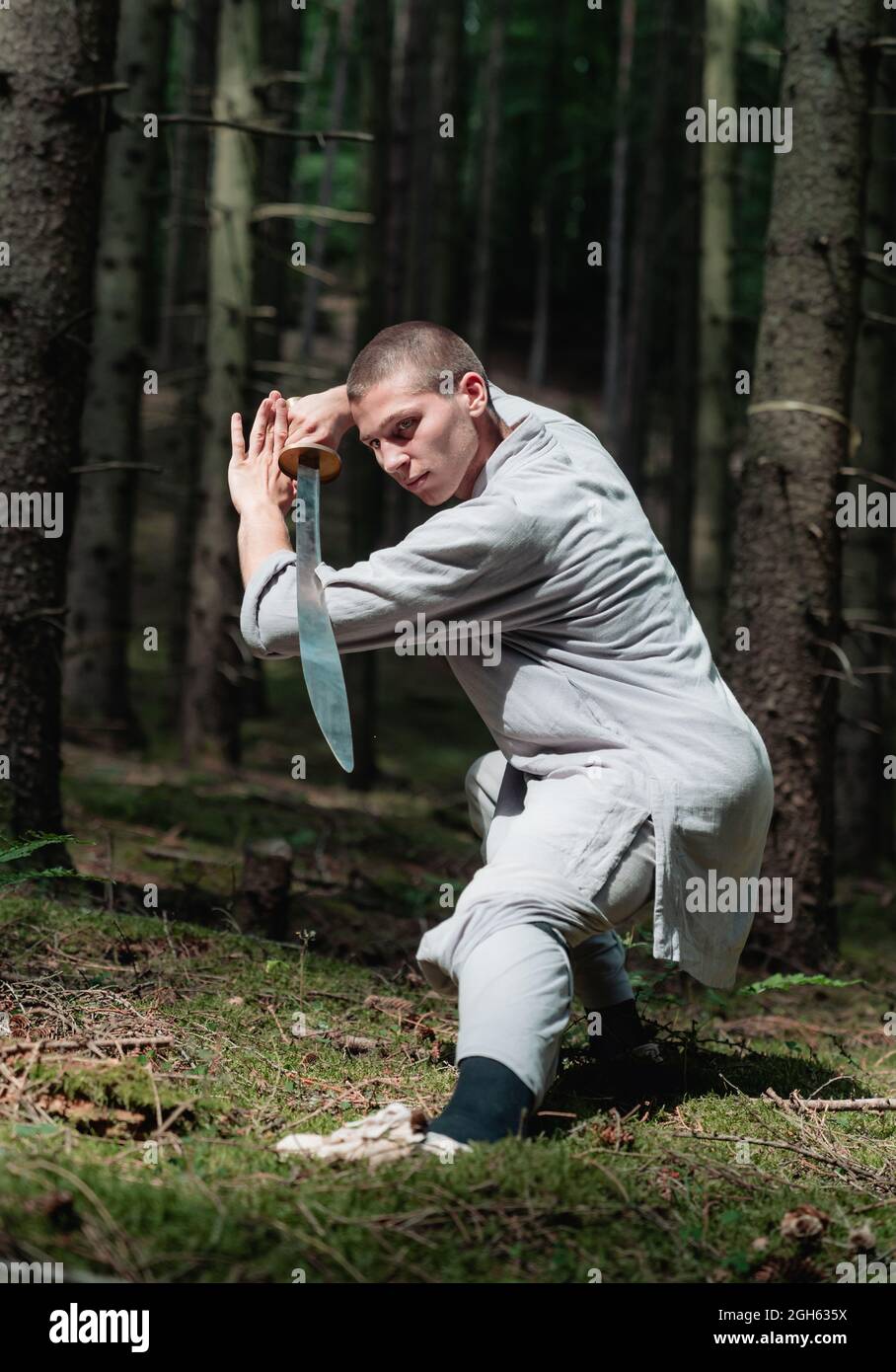 Full body man in traditional clothes practicing sword stance during kung fu training in forest Stock Photo