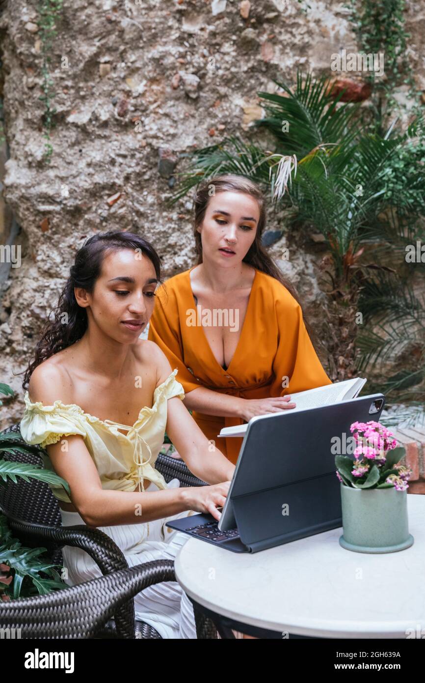 Multiracial female freelancers sitting at table in tropical cafe and working on project while typing on keyboard dock of tablet Stock Photo
