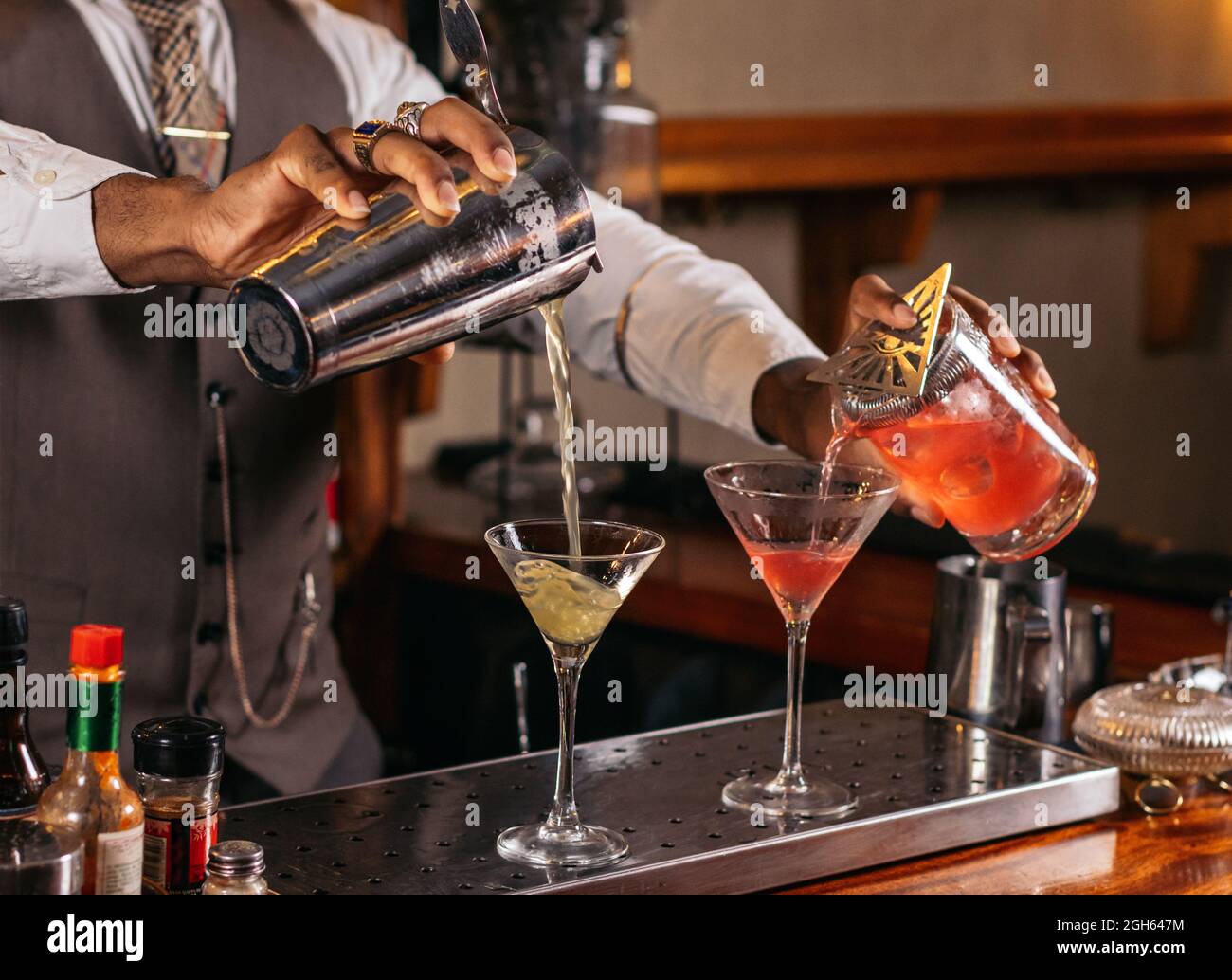 Crop anonymous stylish ethnic male bartender pouring alcoholic drinks from shakers into glasses
