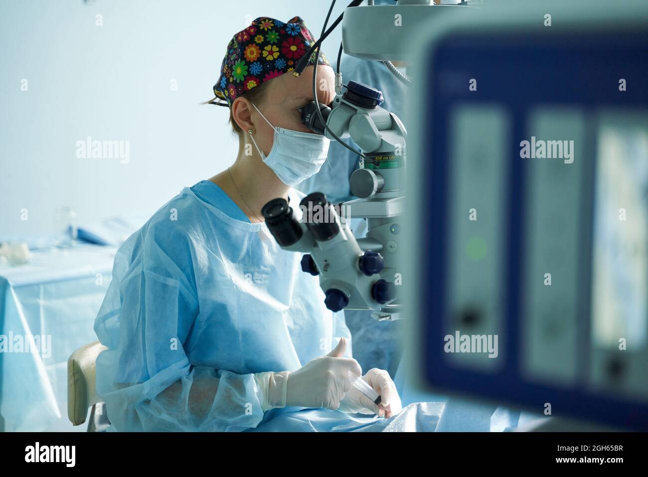 Attentive female doctor in surgical uniform and sterile mask looking through microscope while operating eye of unrecognizable patient in hospital Stock Photo