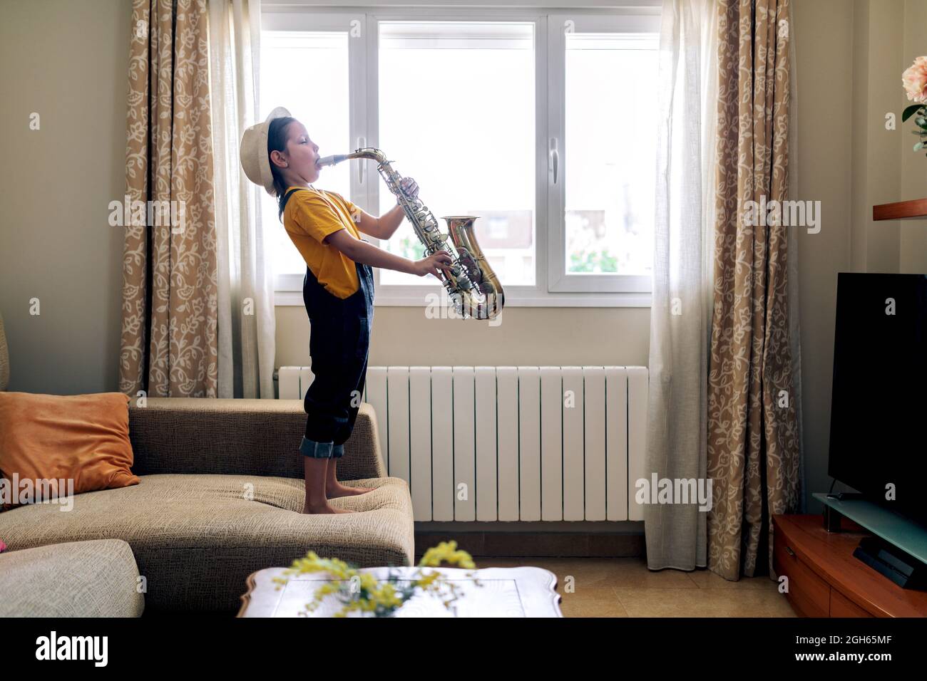 Side view of barefoot child with closed eyes playing saxophone while standing on couch at home in daytime Stock Photo