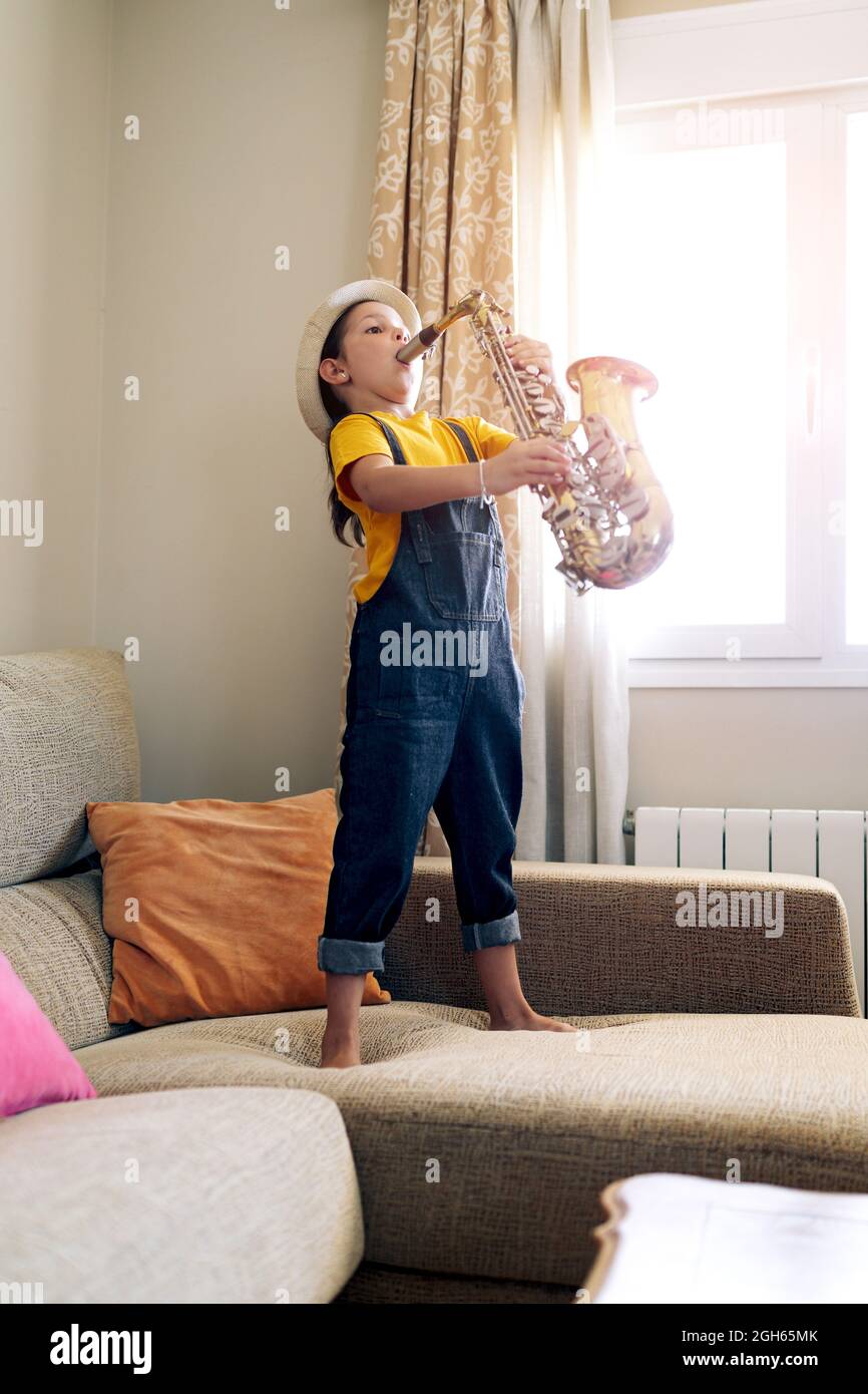 Side view of barefoot child playing saxophone while standing on couch at home in daytime Stock Photo