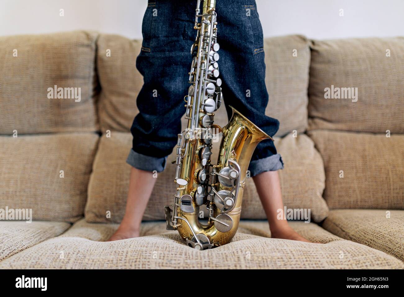 Cropped anonymous thoughtful barefoot child in hat with saxophone standing on sofa against window at home Stock Photo