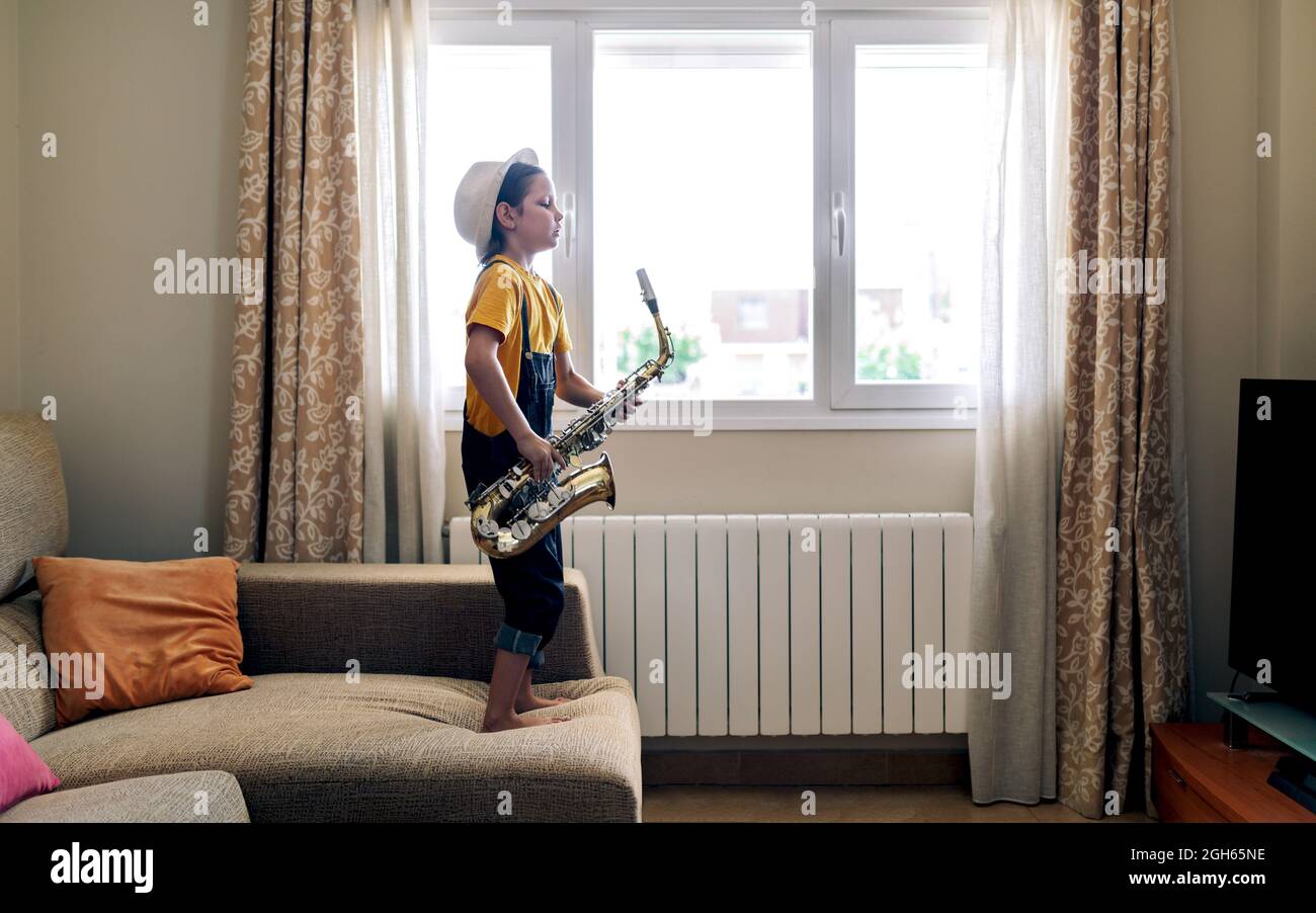 Side view of barefoot child with closed eyes playing saxophone while standing on couch at home in daytime Stock Photo