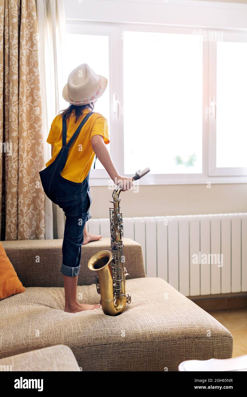 Back view of anonymous thoughtful barefoot child in hat with saxophone standing on sofa against window at home Stock Photo