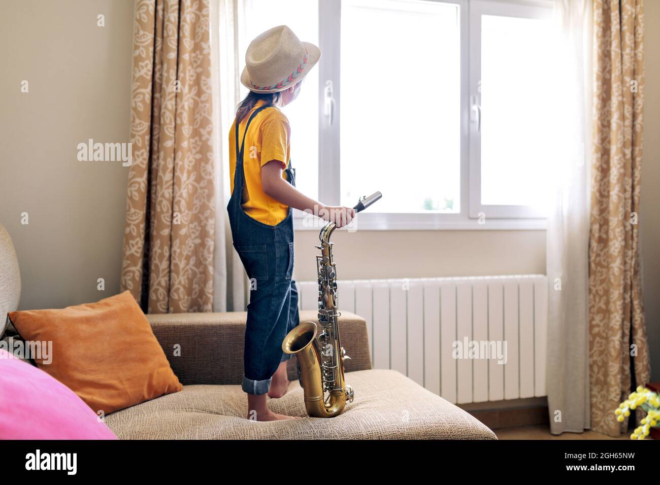 Back view of anonymous thoughtful barefoot child in hat with saxophone standing on sofa against window at home Stock Photo