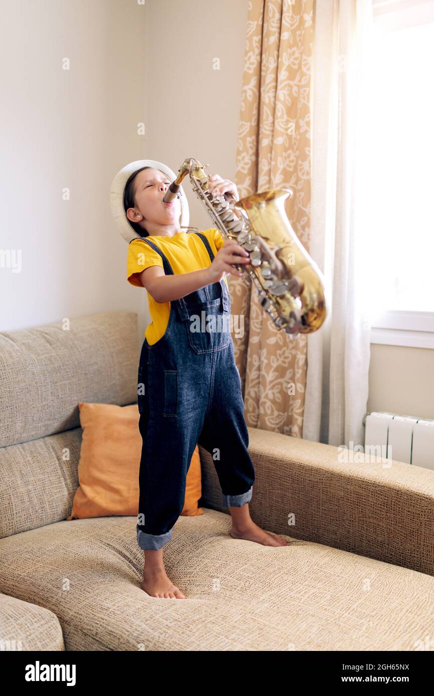 Barefoot child with closed eyes playing saxophone while standing on couch at home in daytime Stock Photo