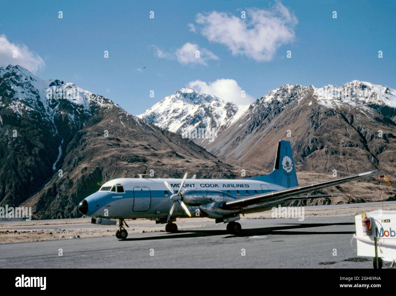 A Hawker Siddeley HS 748 airliner of New Zealand’s Mount Cook Airlines on the tarmac at Queenstown Airport in 1979 with the Southern Alps behind. Mount Cook Airline was a regional airline, founded in 1920, based in Christchurch. It later became a subsidiary of Air New Zealand and in 2019 the brand name was retired. This aircraft is named ‘Aorangi’, a Maori name. On the aircraft tail is an image of the Mount Cook lily (Ranunculus lyallii), the largest buttercup in the world. This image is from an old amateur Kodak colour transparency – a vintage 1970s photograph. Stock Photo