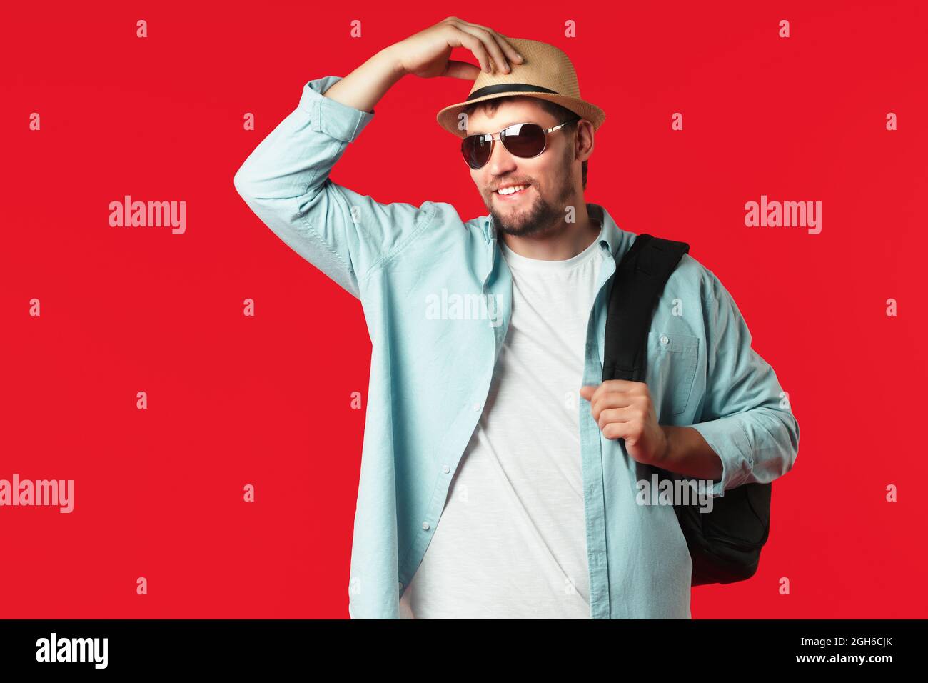 This is a portrait of a man tourist in a hat and a backpack on his shoulder on a red background. Happy guy in casual clothes with sunglasses. Stock Photo
