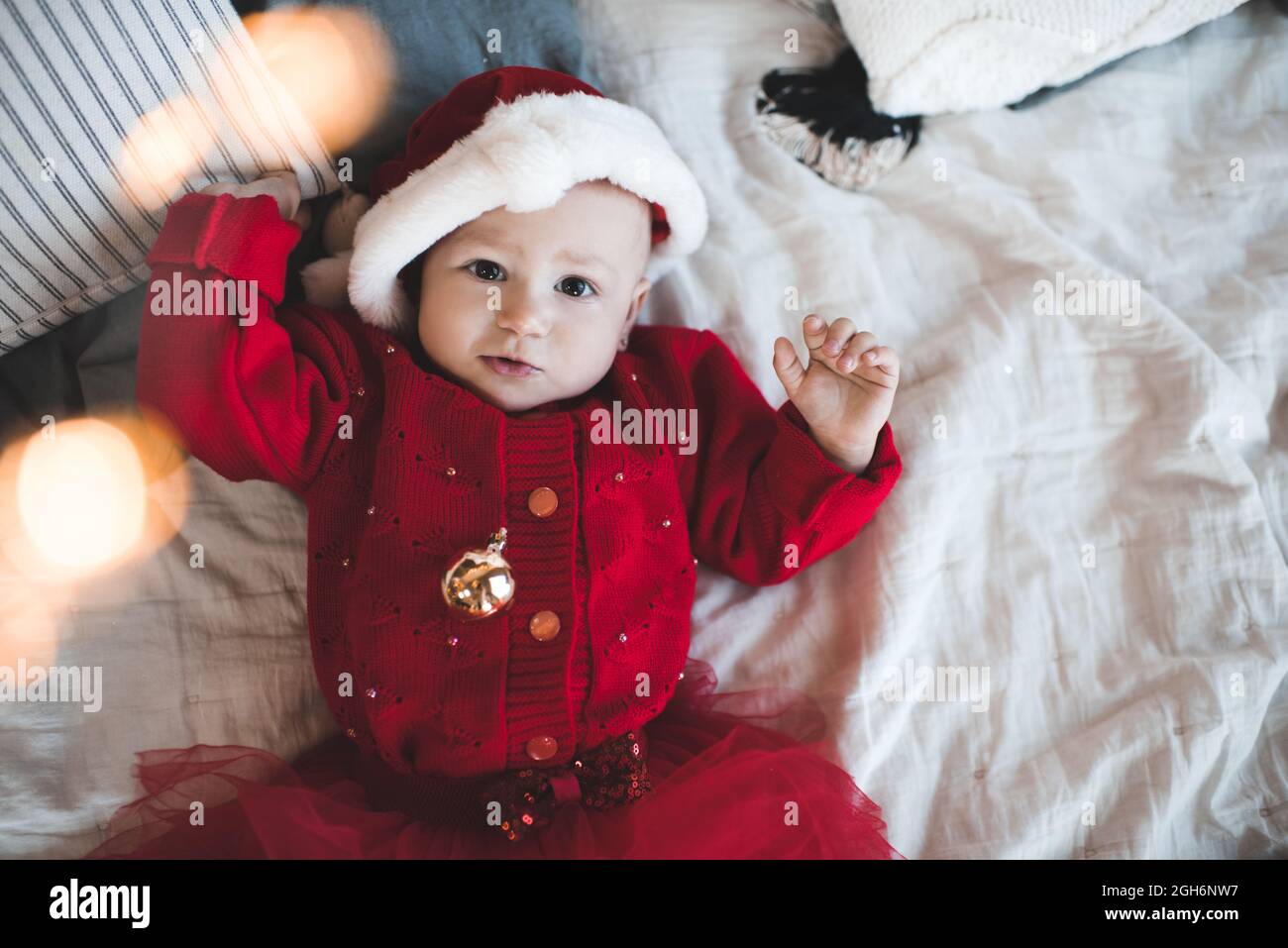 Cute baby girl 1 year old wear red santa claus hat and knit dress lying in bed close up. Smiling