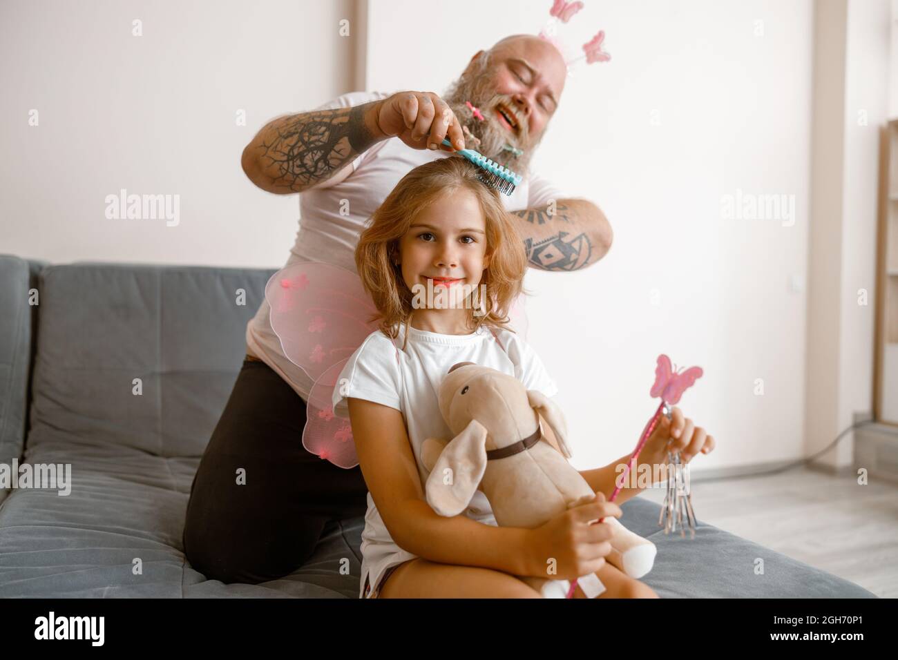 Smiling girl holds toy dog and magic stick while dad brushes her hair in room Stock Photo