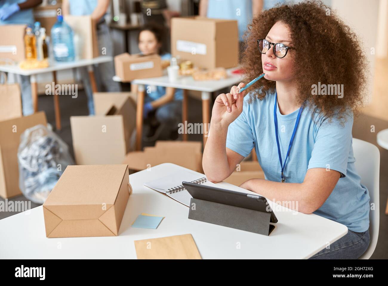 Thoughtful young woman, volunteer in blue uniform looking away, using tablet pc while sitting and working on donation project. Team sorting, packing Stock Photo