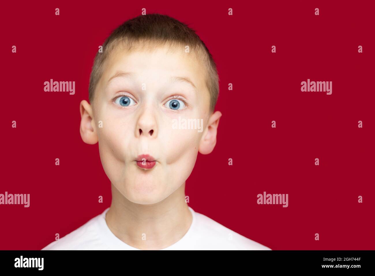 Boy teenager 7-10 in a white t-shirt makes faces, depicting a kiss, with wide open eyes on a red background Stock Photo