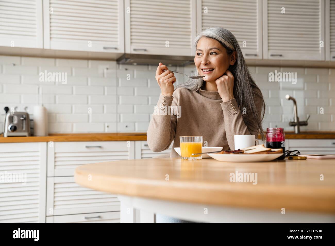Mature grey woman smiling while having breakfast in kitchen at home Stock Photo