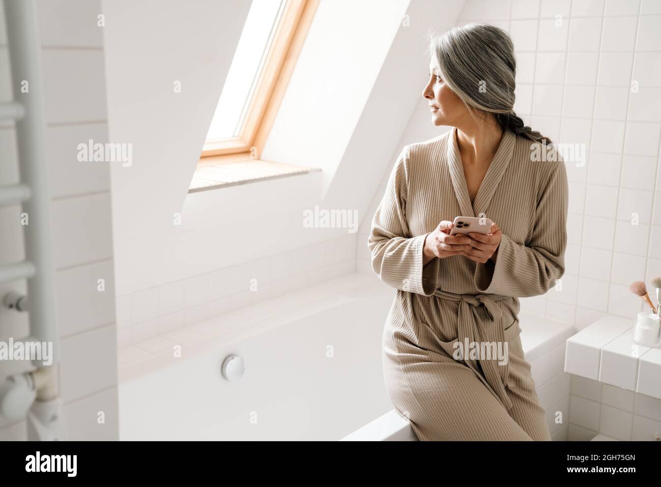 Mature grey woman using mobile phone in bathroom at home Stock Photo