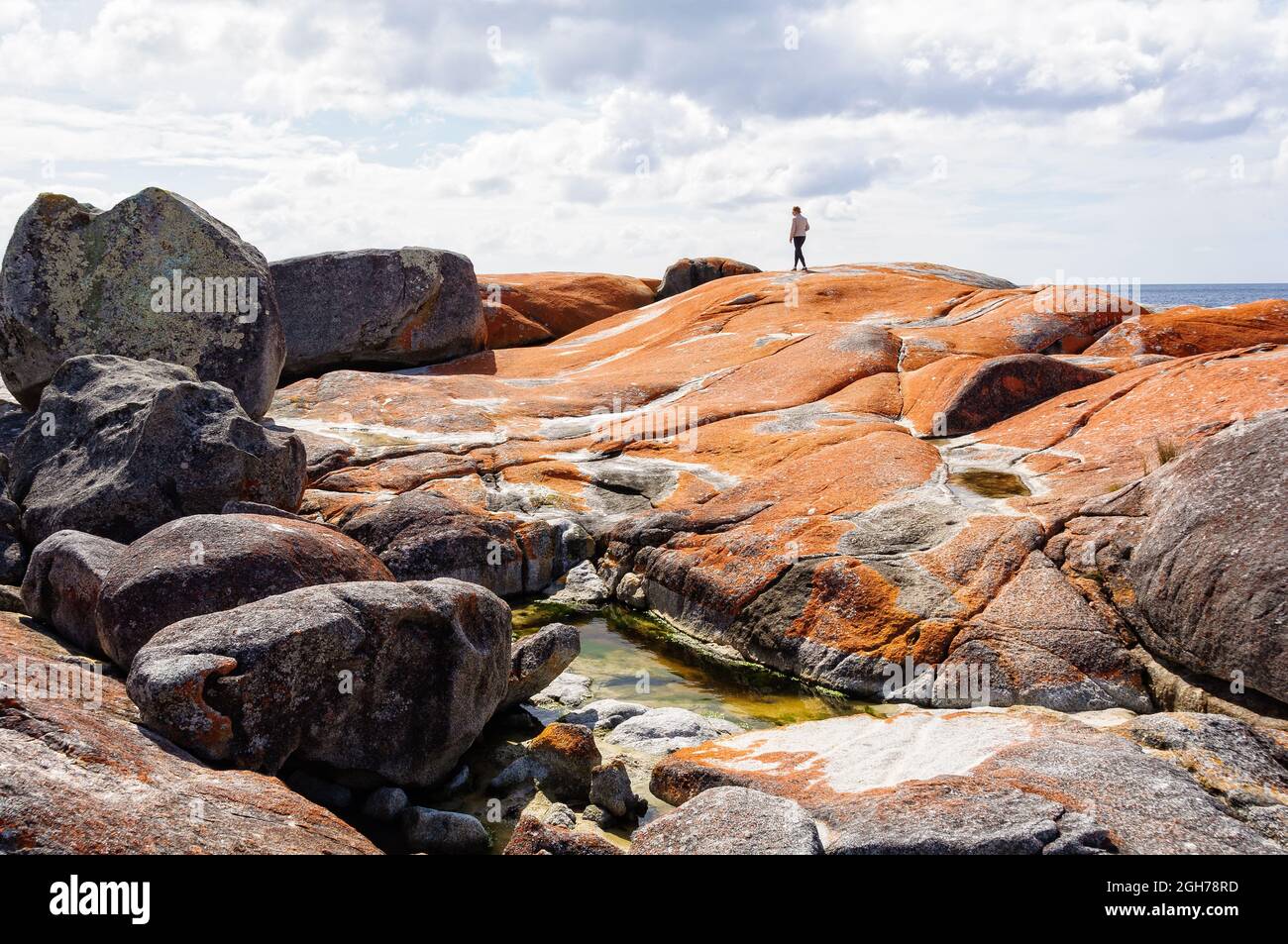 A tourist admires the view from the top of orange lichen-covered granite boulders in Bay of Fires - The Gardens, Tasmania, Australia Stock Photo