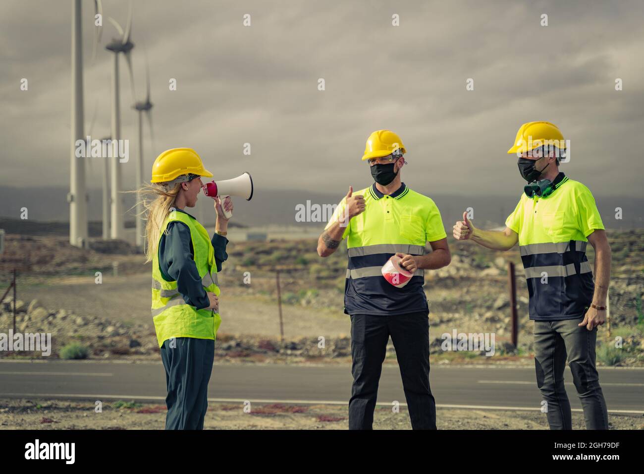 Engineer woman shouts over a megaphone. Skilled workers with protective mask, doing ok sign, thumb up with fingers. Electric central. Stock Photo