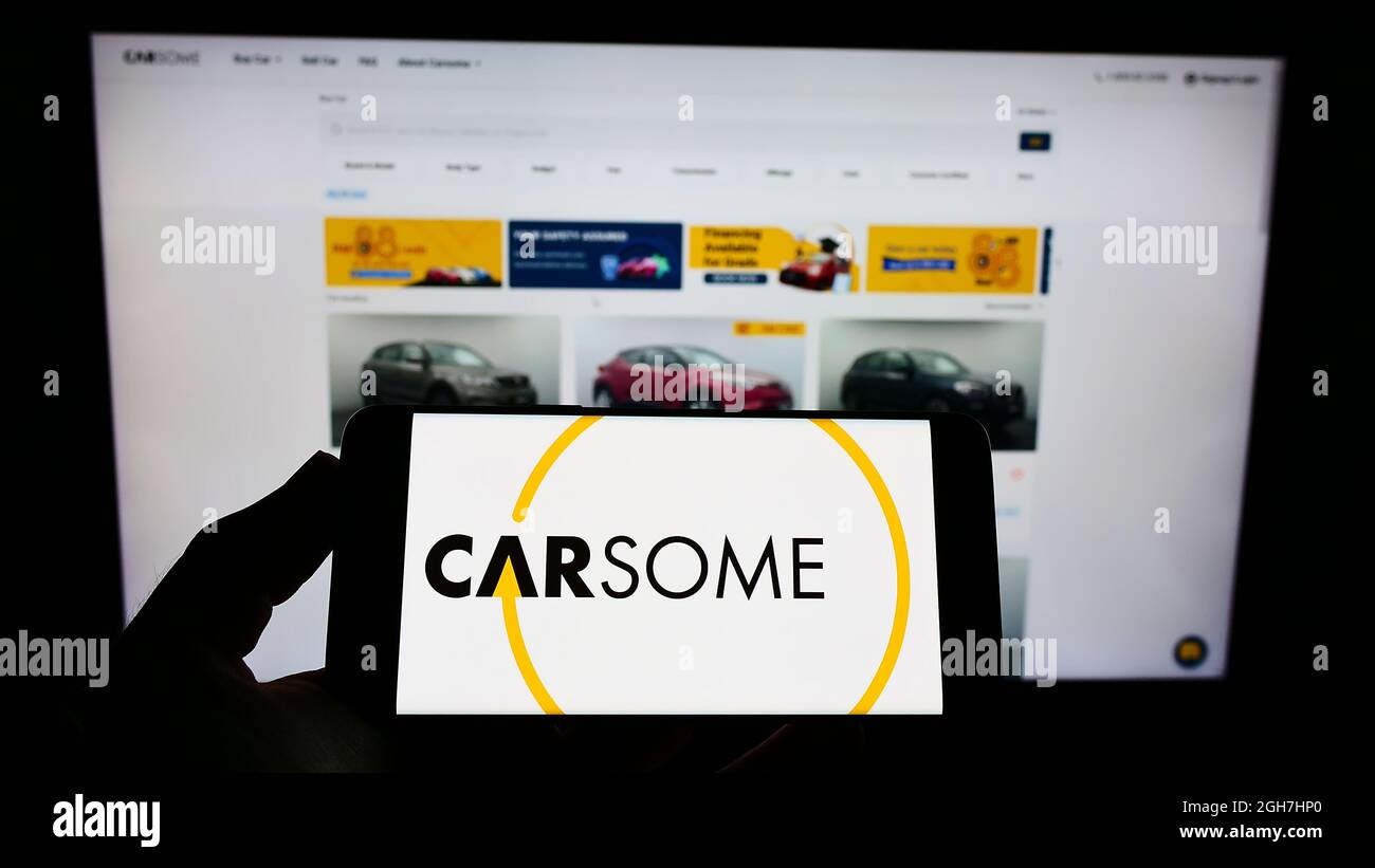 Person holding smartphone with logo of Malaysian used car platform company Carsome Group on screen in front of website. Focus on phone display. Stock Photo