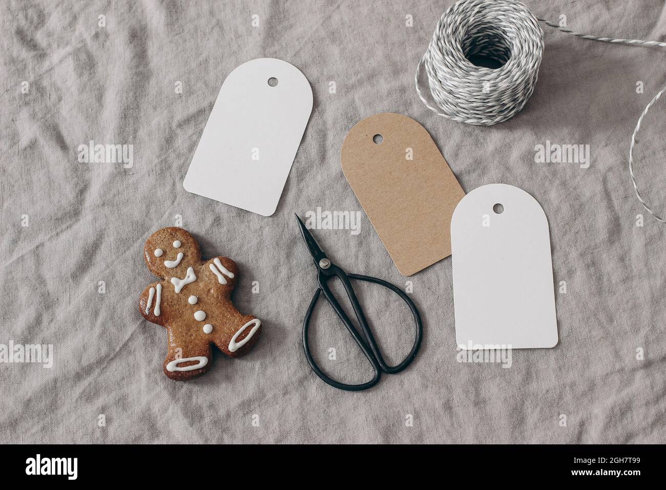 Christmas still life. Blank paper gift tags, labels mockups. Gingerbread cookie, black vintage scissors and decorative rope on beige linen tablecloth Stock Photo