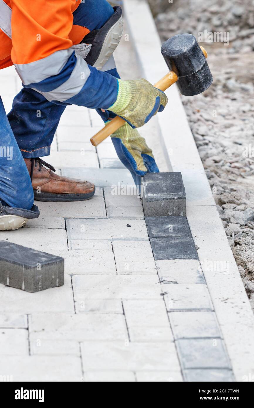 A worker in blue and orange overalls bangs the paving slabs with a rubber mallet. Vertical image, selective focus, copy space. Stock Photo