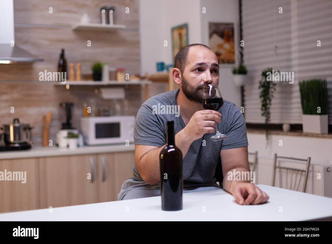 Caucasian man holding glass of wine sitting in kitchen at home. Depressed person drinking liquor, booze, alcoholic beverage alone while feeling intoxicated with bottle of alcohol Stock Photo