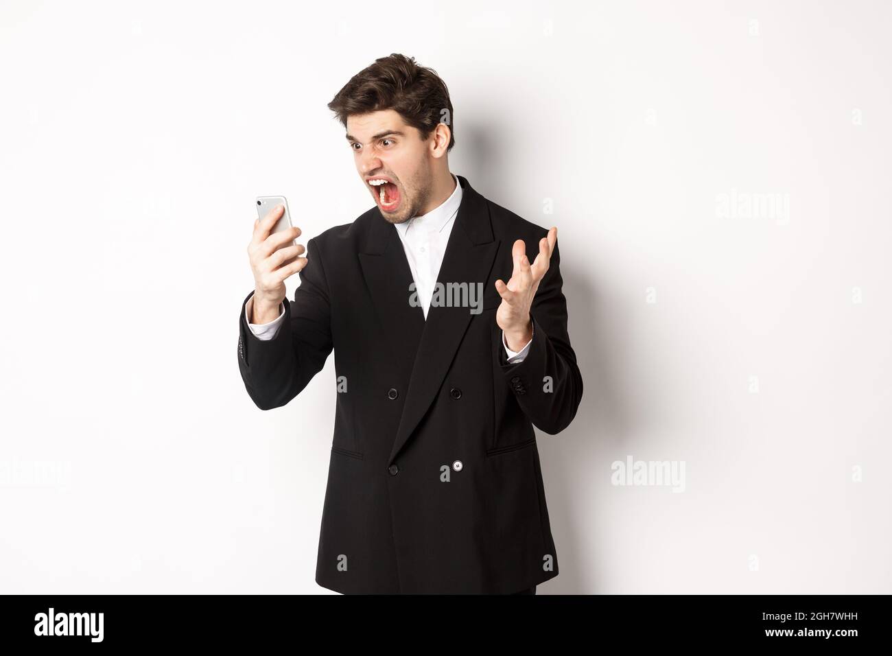 Portrait of angry businessman in black suit yelling at mobile phone, having an argument on video call, standing mad over white background Stock Photo