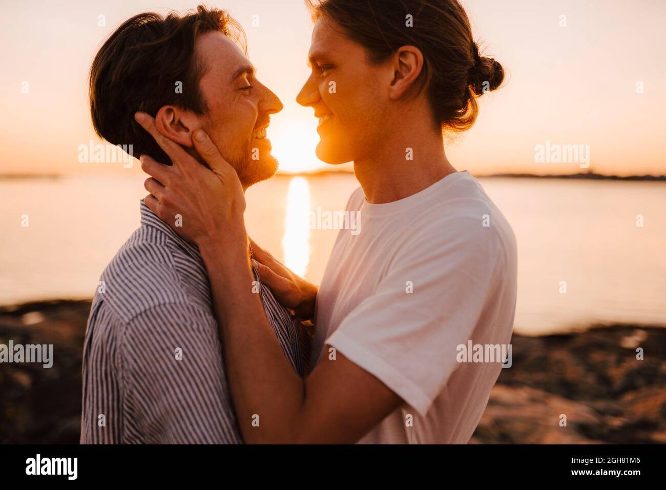 Smiling gay couple standing face to face at lakeshore during sunset Stock Photo